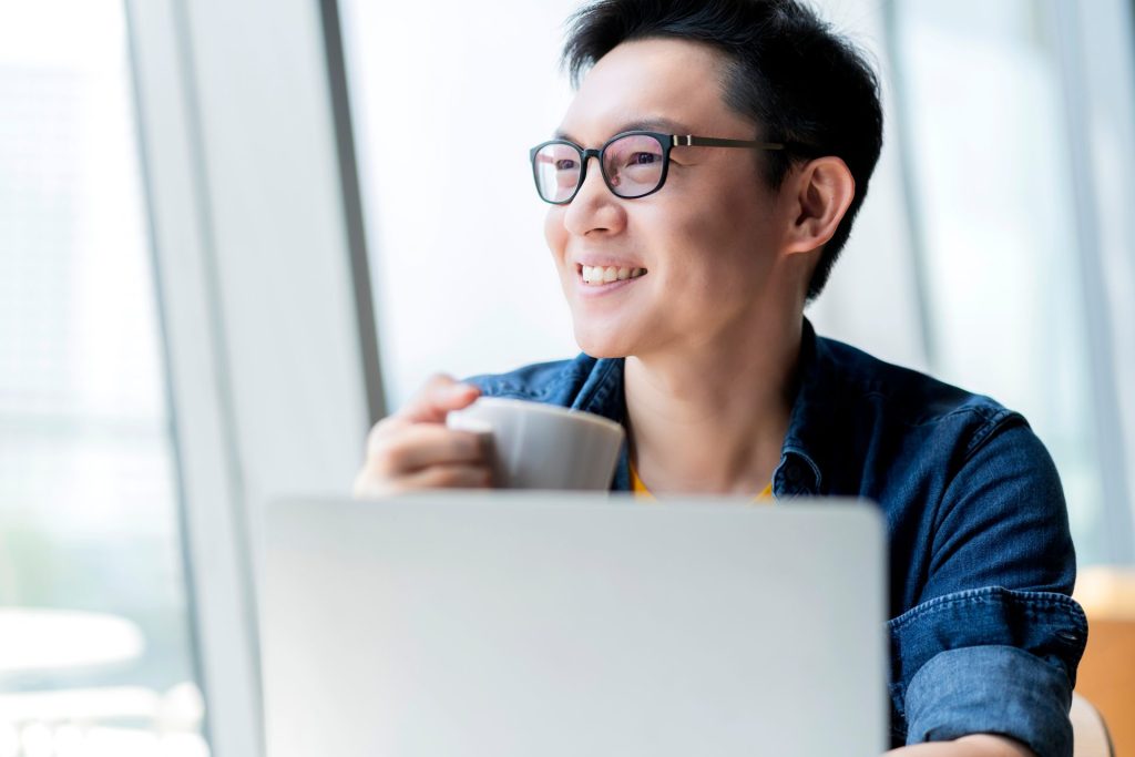 A man with glasses sitting behind his laptop with a cup of coffee